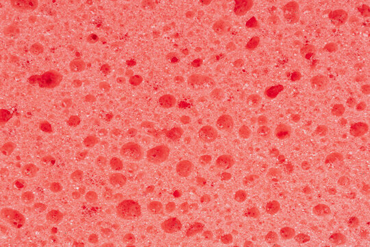 Macro shot of a red foam sponge surface showing a complex network of open cells and polymer fibers. This vibrant porous texture creates deep shadows and 3D relief