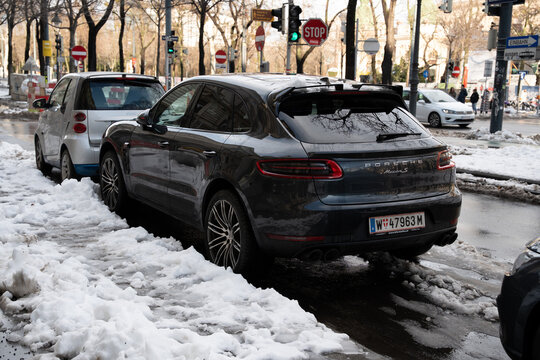 Vienna, Austria- Feb 21, 2026: A Porsche Macan parked beside a small car, partially covered in snow, with traffic lights and street signs in the urban background.