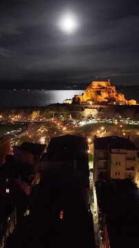Corfu Town night skyline and harbor in Greece. Beautiful aerial view of Kerkyra old town streets, Spianada square, and Ionian sea at nighttime with bright moon, street lights, and urban architecture