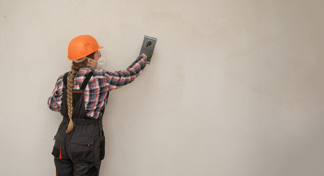 Builder woman in hardhat and protective mask sanding and smoothing walls after plastering for leveling getting it ready for painting during home renovation and construction work. banner.