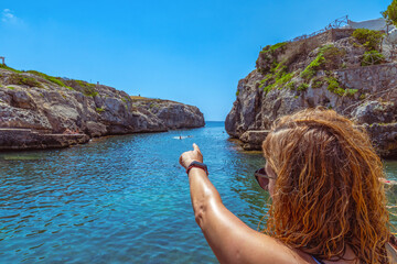 A young, tanned woman points to her friends playing in the water of a beautiful turquoise cove in Menorca, Balearic Islands, Spain.