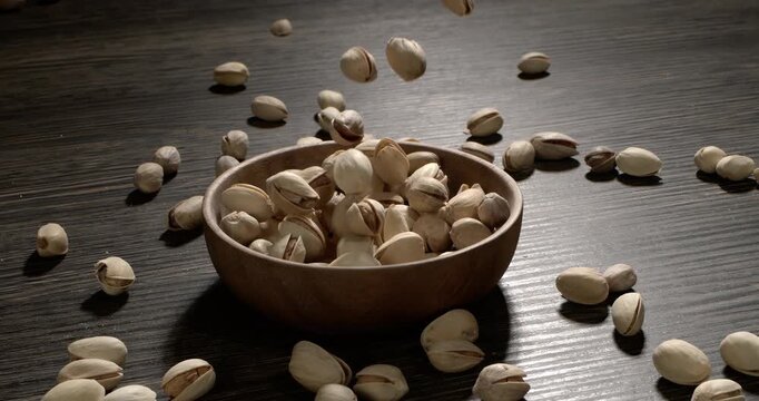 Wooden bowl filled with pistachios on dark wooden table, tasty salted nuts scattered around, healthy snack and natural food concept. Shot on super slow motion camera 1600 fps.