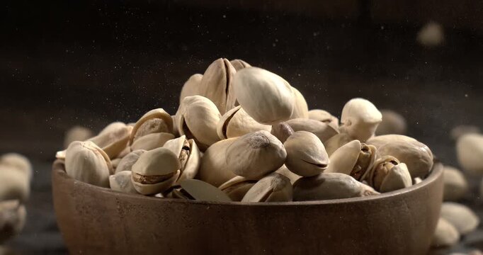 Wooden bowl filled with pistachios on dark wooden table, tasty salted nuts scattered around, healthy snack and natural food concept. Shot on super slow motion camera 1600 fps.