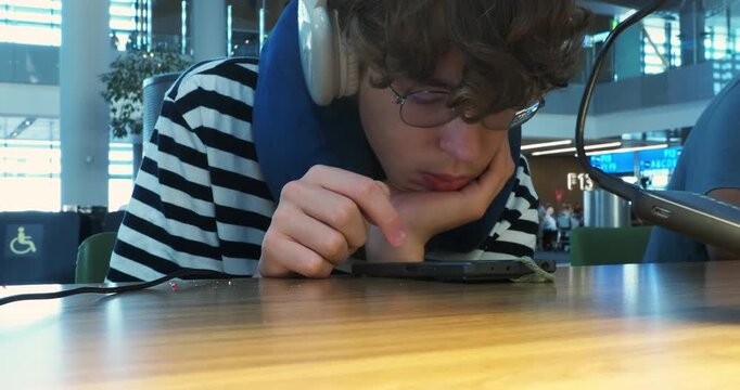 A tired young man with glasses and a travel pillow waits at an airport gate, endlessly scrolling on his smartphone. Close-up shot showing boredom and digital distraction while traveling