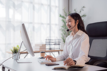 Obraz premium Young Asian woman wearing a headset at a bright office desk, smiling while typing at a computer and providing friendly online customer support and virtual assistance