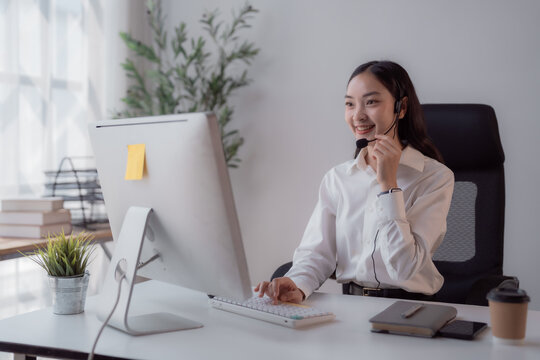 Asian woman smiling and communicating with a client through a headset, providing customer support and telemarketing services from her home office setup
