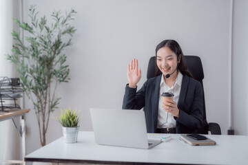 Young Asian businesswoman smiling and waving during a virtual meeting, holding a coffee cup while...