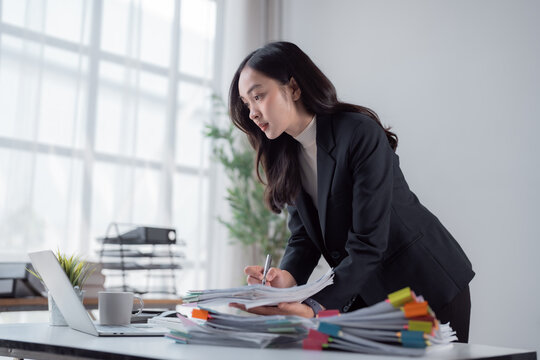 Young Asian businesswoman managing a large pile of documents and files on her desk, carefully reviewing the information while working productively in a modern office environment