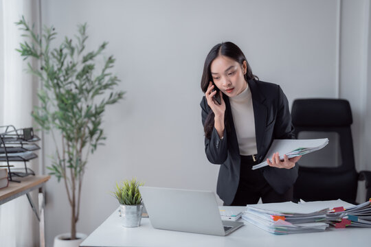 Asian businesswoman managing multiple tasks, talking on a mobile phone while handling a stack of paperwork and using a laptop in a modern office environment