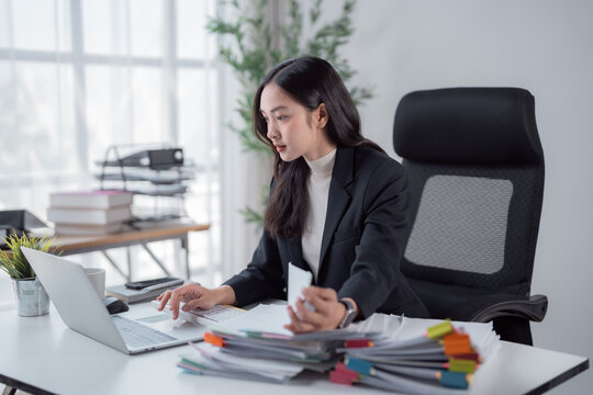 Young professional businesswoman working at an office desk, managing multiple tasks with a laptop and a large stack of categorized paper documents, demonstrating efficiency