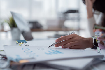 Businesswoman's hand holding a pen, pointing at charts and graphs on financial documents, focusing on data analysis and strategic planning in a professional office environment © MINAE