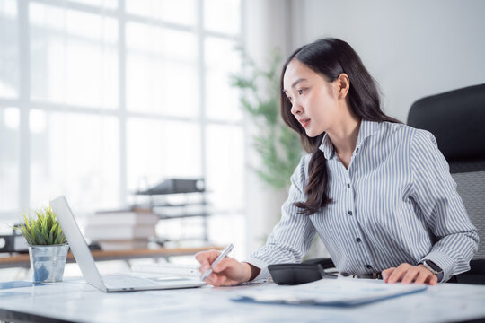 Asian businesswoman focused on her laptop while taking notes and managing documents at a tidy desk in a bright, modern office, embodying professionalism and productivity