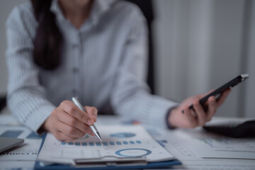 Fototapeta premium Businesswoman's hands reviewing financial report graphs and charts on a clipboard, using a pen to indicate figures while holding a smartphone for additional data