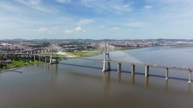 Aerial drone footage flying towards the majestic Vasco da Gama Bridge over the Tagus River in Lisbon, Portugal. Shows modern architecture, city skyline, and scenic landscape on a sunny day