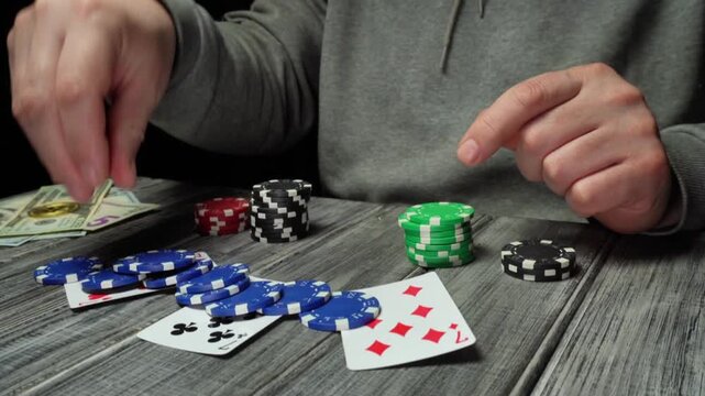 The hands of a male gambler in a gray sweatshirt carefully stack chips on three playing cards on a wooden table, cash and additional chips are visible in the background