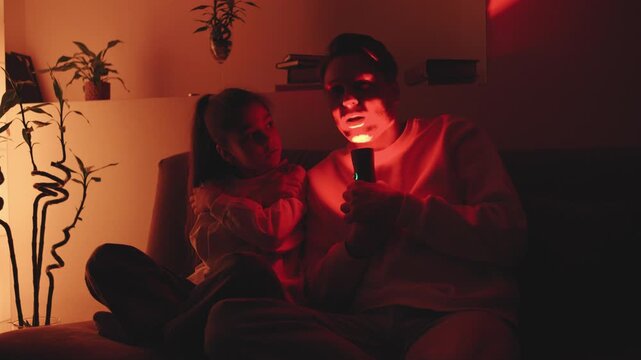 A father sits on a couch in a living room with a child. They are using a flashlight to tell stories. The room has warm light and plants in the background.