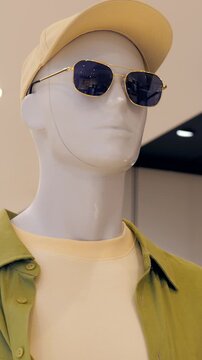 Beige cap and sunglasses styling on mannequin with green overshirt and yellow tee, summer commuter look staged by visual stylist, gold frames catching store lights, relaxed urban presentation