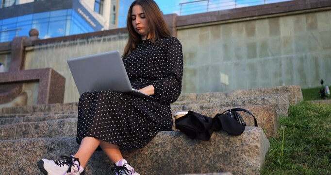 Female on steps researching outdoors. Woman in polkadot dress working on laptop exterior at dusk. Caucasian woman in polkadot dress focused on studying outdoors near building during dusk