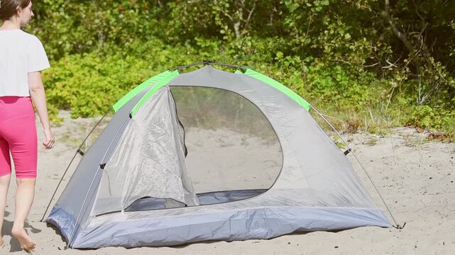 Woman inspecting gray and green tent tautness on sandy beach with green foliage background