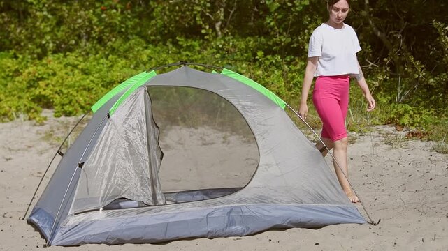 Woman inspecting and adjusting tent tautness on sandy outdoor campsite with green forest background