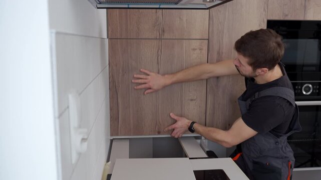 Man in overalls installing wooden kitchen cabinet door in modern apartment interior. Concept of home renovation self made furniture assembly and repair service