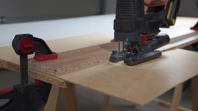 Close up of electric jigsaw cutting MDF board fixed with clamps on workbench in workshop. Concept of woodworking DIY repair and construction process