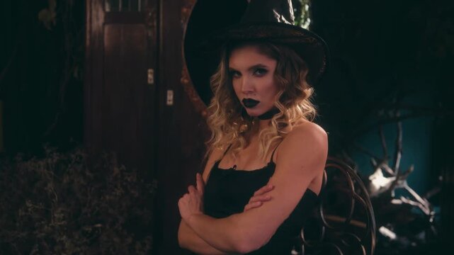 Beautiful woman in a black witch costume with a pointy hat and dark makeup posing with her arms crossed in a spooky decorated room, looking seductively at the camera for halloween