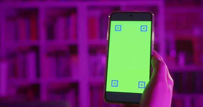 Woman holding a smartphone with a green screen at home in the night.