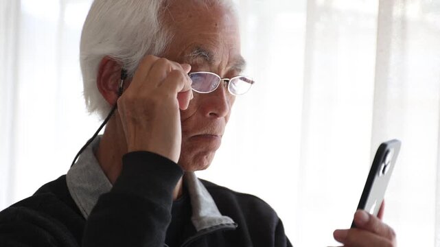 Slow motion of senior Japanese man with glasses using smartphone at home, cinematic lifestyle, silver generation, farsightedness, focused expression, elderly, 4K 30p, natural light.