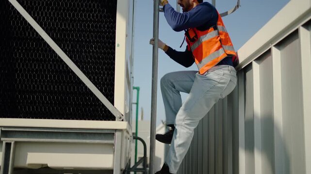 Professional technician wearing safety gear and a yellow hard hat climbs a caged metal ladder to inspect an industrial cooling tower on a sunny rooftop, focusing on facility maintenance and safety.