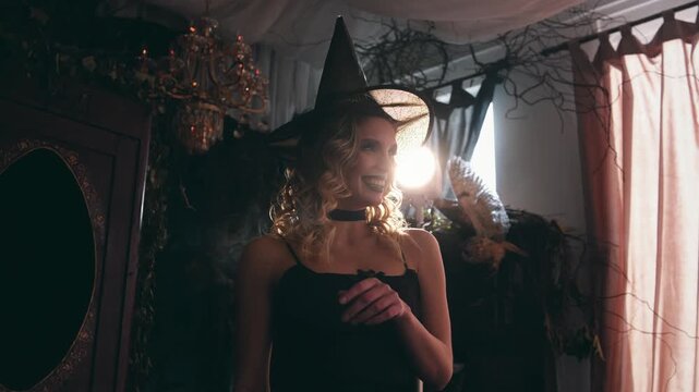 Joyful blonde woman wearing a black witch hat and dress laughing and dancing in a dark, spooky decorated room during a halloween party, with backlight creating a magical atmosphere