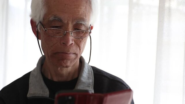 Slow motion of senior Japanese man with glasses using smartphone at home, cinematic lifestyle, silver generation, farsightedness, focused expression, elderly, 4K 30p, natural light.