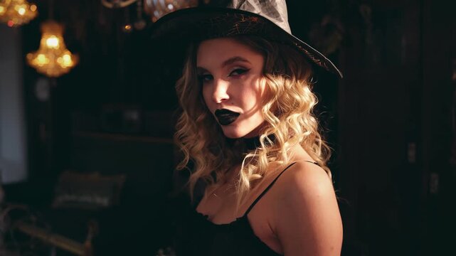Seductive blonde woman wearing a witch hat and black lipstick for halloween, looking at the camera with a mysterious expression in a dark, atmospheric room with warm lights in the background