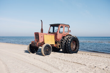 weathered, vintage red tractor stands parked on a sandy beach directly facing the calm blue ocean © Jarama