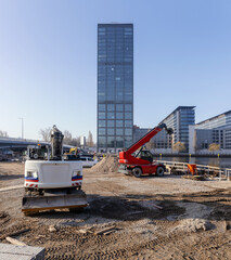 A busy urban construction site in the foreground with a mini excavator and red telescopic handler crane, set against a tall modern glass office tower and clear blue sky in a city environment