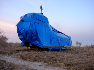 A large vessel completely wrapped in a bright blue tarpaulin stands elevated on supports in a dry coastal meadow, with a hazy sea visible in the background