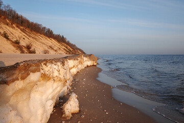 dramatic coastal scene featuring a thick ledge of ice and frozen sand along a beach, with steep sandy cliffs in the background