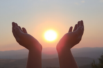 Fototapeta premium Two hands gently cupping a bright sun at dawn with soft golden light and a serene sky