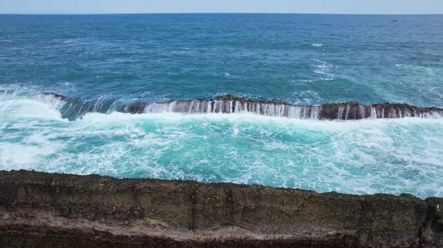Aerial drone footage of Tanjung Layar pointy rock bareer formation, a flat rocky beach, blue sea, with many waves hitting the reef, streams like waterfalls, in Sawarna beach, Banten, Java, Indonesia