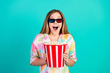 Young woman wearing 3D glasses with popcorn, expressing excitement, turquoise background