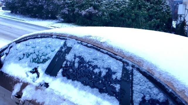 A car is partially covered with a layer of white snow, parked near a road with some dry grass in the background, indicating a cold winter day. A car's windshield and hood are blanketed in soft white s