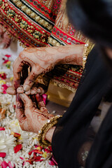 Woman's mehndi adorned hands place toe ring on another woman's foot in a cultural ceremony on a bed of white rice and flower petals