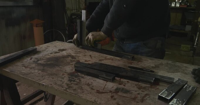 Professional metalworker using a try square to align two pieces of rectangular steel tubing at a perfect right angle on a workbench before welding, ensuring precision in a rustic workshop.
