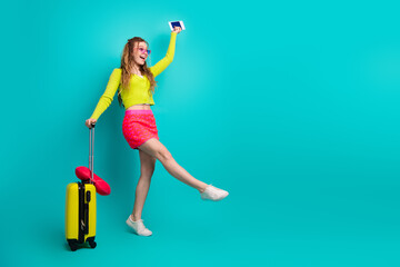 Fototapeta na wymiar Stylish young woman holding passport and luggage poses in trendy outfit with vibrant teal background for travel theme