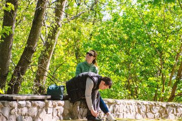 Fototapeta premium Young Couple Relaxing Outdoors in Sunny Spring Park; Man Tying Shoelace While Woman Enjoys Nature