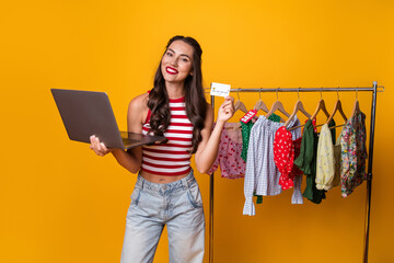 Young woman with laptop and credit card shopping for clothes on a bright yellow background