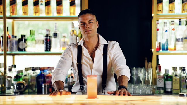 Charismatic male bartender standing behind a well stocked bar counter, presenting a refreshing cocktail with a confident look