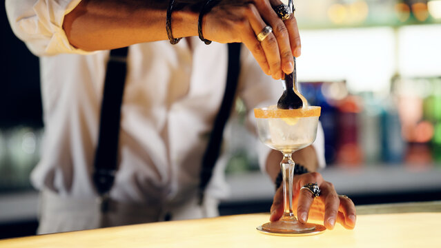 Bartender's hands preparing a beverage, adding ingredients to a cocktail glass with a sugar rim at a bar counter