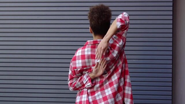 African-American woman stretches her arms behind her back against urban wall, girl practices yoga and fitness and warms up her ligaments and tendons outdoors