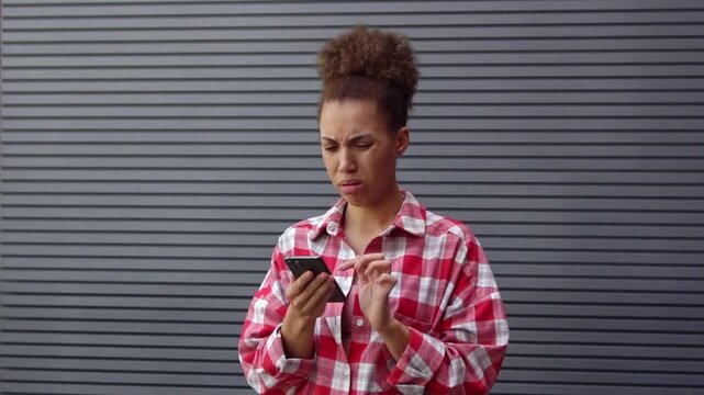 African American woman using smartphone and expressing disgust in front of urban wall, girl will watch disgusting and unpleasant online content outdoors